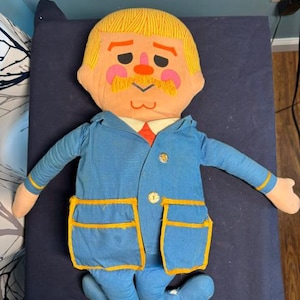 May include: A plush doll featuring a character with yellow hair, a mustache, and pink cheeks. The doll wears a blue jacket with yellow trim, two large pockets, and two gold-colored buttons, over a white shirt with a red tie and blue pants.