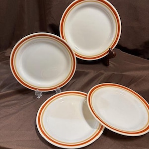 Puede incluir: Un juego de cuatro platos de cerámica blanca con un diseño retro. Cada plato presenta una banda ancha naranja y una fina banda beige alrededor del borde. Los platos están colocados sobre una superficie marrón.