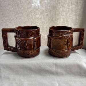 May include: Two brown ceramic mugs with square handles. The mugs have a detailed, embossed design around the body, featuring a belt or strap motif. The mugs are on a white surface.