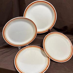 Puede incluir: Cuatro platos de cerámica blanca con un borde marrón y beige. Los platos están dispuestos sobre una superficie marrón. Los platos son de diferentes tamaños y formas, incluyendo redondos y ovalados.