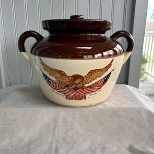 May include: A brown and cream-colored ceramic crock with a lid and two handles. The crock features an eagle design with red, white, and blue accents. The text "The Spirit of America" is printed below the eagle.