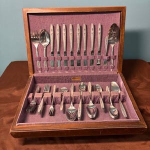Peut inclure: Un service de couverts vintage présenté dans un coffret en bois avec une doublure en velours violet. L'ensemble comprend des fourchettes, des cuillères, des couteaux et des ustensiles de service. Les couverts ont un motif floral décoratif sur les manches.
