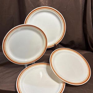 Puede incluir: Cuatro platos de cerámica blanca con un diseño de borde marrón y naranja. Los platos están dispuestos sobre una superficie marrón. Los platos son de diferentes tamaños y formas.