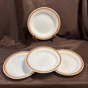 Puede incluir: Cuatro platos de cerámica blanca con un diseño de borde marrón y naranja. Un plato está sobre un soporte de madera. Los platos están dispuestos sobre una superficie marrón.