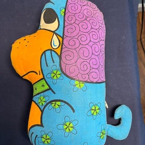 May include: A stuffed fabric dog toy featuring a bright orange face with a single blue teardrop, a large purple ear with a black spiral pattern, and a blue body decorated with black-outlined green flower motifs and a small curled tail.