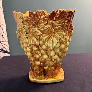 May include: A ceramic vase featuring a three-dimensional relief of grape clusters and vine leaves. The piece is finished in a glossy glaze with shades of olive green, light green, and brown. It has a wide, scalloped rim and a rectangular base.