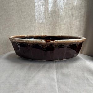 Puede incluir: Una fuente ovalada de cerámica marrón con borde festoneado. La fuente tiene un acabado brillante y un color marrón oscuro con un borde marrón más claro. La fuente es probablemente vintage y adecuada para hornear.