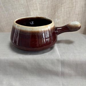 Puede incluir: Un cuenco de sopa de cerámica marrón con asa. El cuenco tiene un exterior marrón oscuro con un borde marrón claro y crema. El interior es oscuro. El asa es de color marrón claro. El cuenco está sobre una superficie blanca.