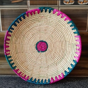 May include: A round woven basket with a pink and teal trim. The basket is made of natural materials and has a circular design in the centre.