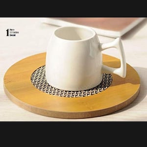 Puede incluir: Una taza de cerámica blanca con un asa única se encuentra sobre un posavasos redondo de madera. El posavasos tiene un centro oscuro estampado y un borde de madera clara. El texto de la imagen dice "1 Pieza 14 cm Redondo".