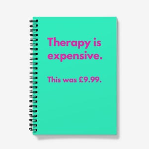May include: A teal spiral-bound notebook with the text "Therapy is expensive." in pink. Below, the text "This was £9.99." is displayed. The notebook has a black metal spiral binding.