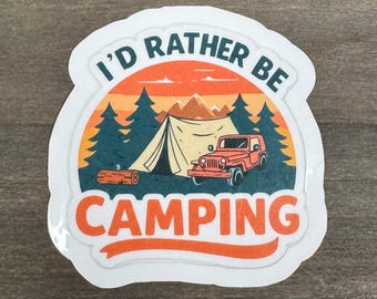 May include: A sticker with a camping theme. The design features a tent, a red jeep, trees, and the text "I'd Rather Be Camping" in a white and orange font. The background has an orange and blue color scheme.