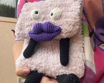 May include: A whimsical, plush, light pink crossbody bag with a cartoonish face. Features large white crocheted eyes, a purple crocheted mouth, and black crocheted legs. The bag has a black chain strap.