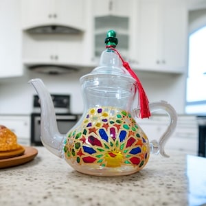Peut inclure: Une théière en verre transparent avec un motif coloré de vitrail sur la moitié inférieure. La théière a un bec courbé, une anse et un couvercle avec un bouton vert et un gland rouge. La théière est sur un comptoir.