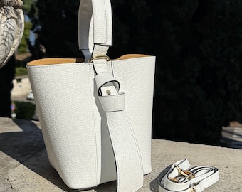 May include: A white bucket bag with a top handle and a detachable shoulder strap. The bag has a textured exterior and gold-colored hardware. The interior lining is a light brown color. The bag is sitting on a stone surface.