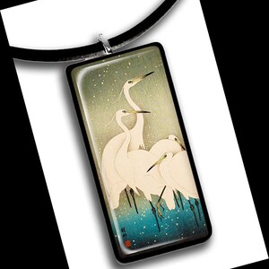 Könnte beinhalten: Eine rechteckige Glas-Anhängerkette mit einem schwarzen Band. Der Anhänger zeigt ein japanisches Gemälde von vier weißen Reihern, die in einer schneebedeckten Landschaft mit blauem Hintergrund stehen.