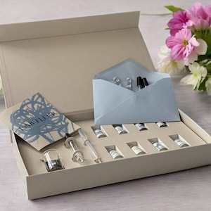 Op de afbeelding: Een beige doos met flesjes, een pipet en een kleine beker. Een lichtblauwe envelop bevat meer flesjes en pennen. Een kaart met de tekst "LAB FLEER" is ook inbegrepen. Roze en witte bloemen staan op de achtergrond.