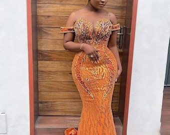 Könnte beinhalten: Ein orangefarbenes, schulterfreies Abendkleid mit aufwendiger Perlenarbeit und einer Meerjungfrauen-Silhouette. Das Kleid hat einen tiefen Ausschnitt und eine fließende Schleppe. Ein passendes Kopftuch vervollständigt das Ensemble. Das Kleid ist für besondere Anlässe geeignet.