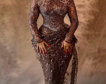 Op de afbeelding: Een bruine, met pailletten bezette avondjurk met lange mouwen en een vierkante halslijn. De jurk heeft ingewikkelde details rond de halslijn en mouwen. Een bijpassende hoofddoek en sieraden maken het elegante ensemble compleet. De jurk is vloerlang.