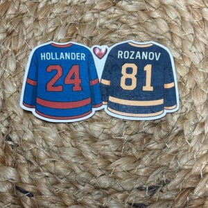 Könnte beinhalten: Zwei Aufkleber in Form von Hockeytrikots. Einer ist blau mit dem Namen "HOLLANDER" und der Nummer "24" in Rot. Der andere ist marineblau mit "ROZANOV" und "81" in Orange. Ein rotes Herz befindet sich dazwischen.