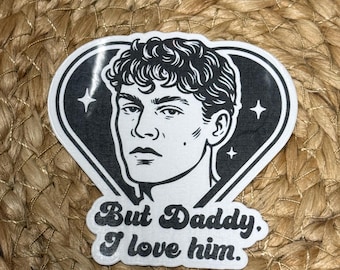 Op de afbeelding: Zwart-witte sticker met een portret van een man in een hartvorm en de tekst "But Daddy, I love him". De sticker is geplaatst op een geweven, beige achtergrond. Close-up.