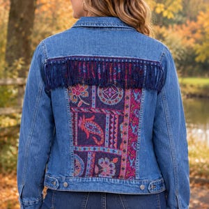 May include: A blue denim jacket featuring a back panel with a colourful paisley and floral pattern in shades of purple, pink, and blue. The jacket includes a horizontal row of dark blue fringe trim across the upper back and metal buttons.