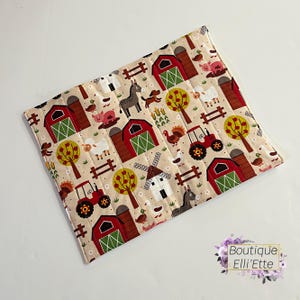 May include: A rectangular, quilted farm-themed placemat. The beige fabric is patterned with red barns, green-roofed silos, tractors, trees, and farm animals. The placemat has a stitched border and a floral logo in the bottom right corner.