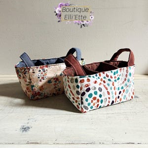 May include: Two fabric storage baskets with handles. One basket features a floral pattern, the other has a white background with colorful abstract shapes. The baskets have interior dividers and are labeled "Boutique Elli'Ette".