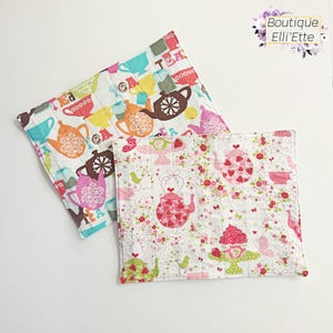 May include: Two square quilted pot holders. One features colorful teacups and teapots, the other has a floral design with teapots, birds, and strawberries. The pot holders are white with colorful accents.