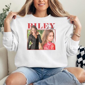 May include: White sweatshirt featuring a graphic of a woman with the text "RILEY Reed" in red. The graphic includes two images of the woman in a black dress and a red patterned top. The sweatshirt is made of a soft material.