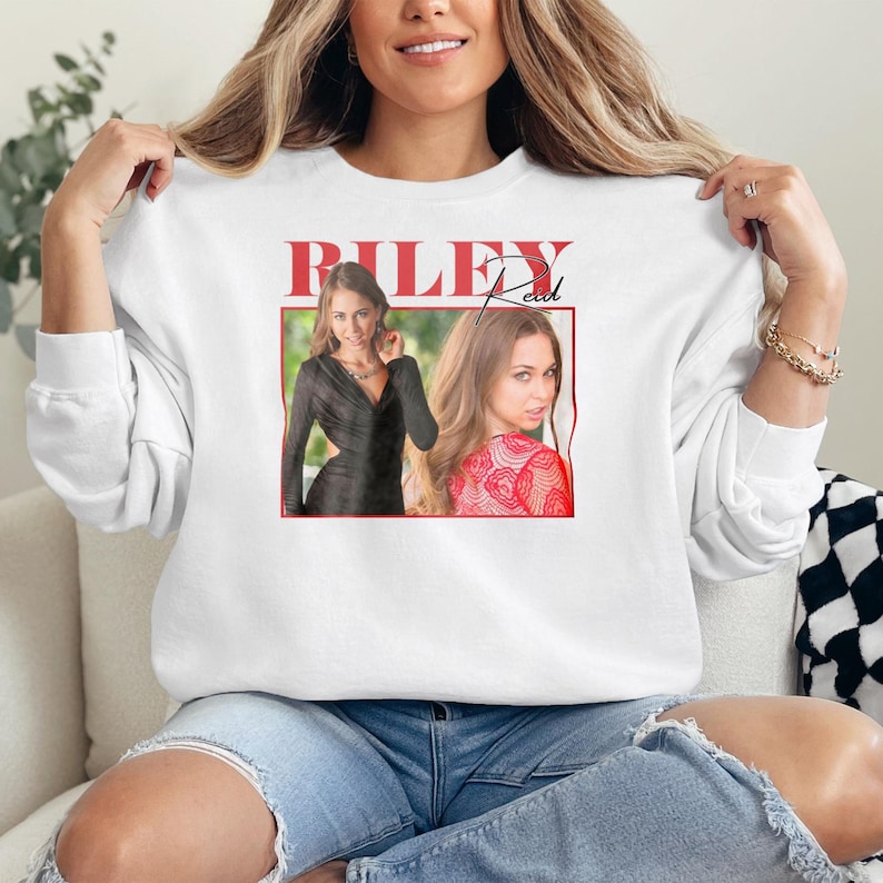 May include: White sweatshirt featuring a graphic of a woman with the text "RILEY Reed" in red. The graphic includes two images of the woman in a black dress and a red patterned top. The sweatshirt is made of a soft material.