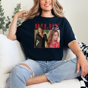 May include: Black t-shirt featuring a graphic of a woman in a black dress and a red patterned top. The name "Riley Reid" is printed in red above the images. The shirt is worn by a person.