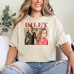 May include: Beige t-shirt featuring a graphic of a woman with the name "RILEY Reid" in red text. The graphic includes two images of the woman. The shirt is a casual style.
