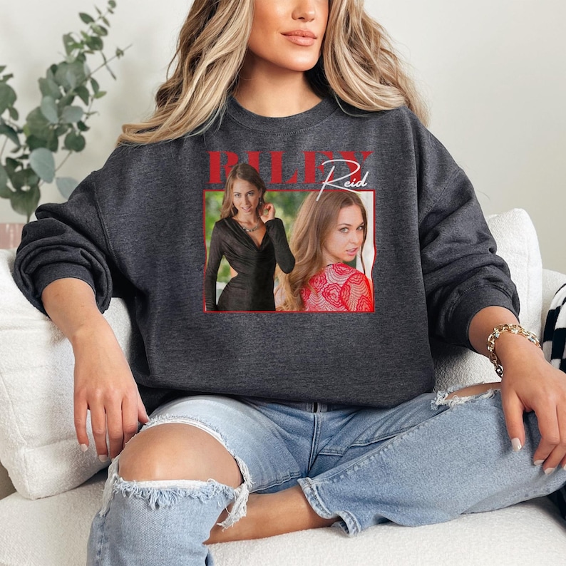 May include: Dark gray sweatshirt featuring a framed image of a woman with the name "RILEY Reid" in red letters. The image within the frame shows two photos of the woman. The sweatshirt is a casual style.