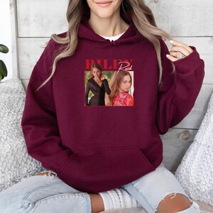 May include: A maroon hoodie with a front pocket and a white undershirt peeking out. The hoodie features a rectangular graphic with the name "RILEY Reid" in red, along with two images of a woman.