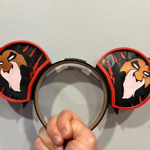 May include: Black and red mouse ear headband featuring a character design. The ears have a black background with a red border and a character face. The headband is held by a hand.