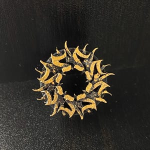 May include: A gold-colored metal ring with a circular design featuring intricate, stylized patterns. The ring is on a black background.