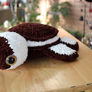 May include: A handmade crochet turtle plushie in shades of brown and white. The turtle has large, expressive eyes with gold accents. The shell and flippers are brown with white trim, and the head is white with brown details.