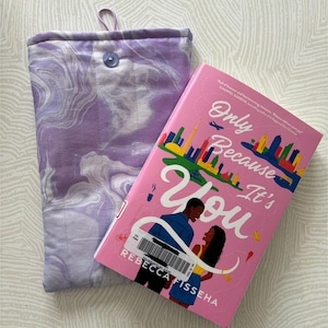 May include: A lilac and white marbled fabric book sleeve with a button closure next to a pink book titled "Only Because It's You" by Rebecca Fisseha. The book cover features a couple and a city skyline.