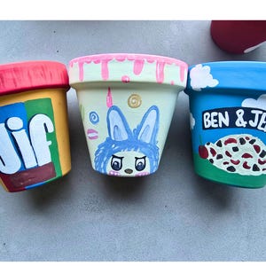 May include: Three hand-painted terracotta flower pots. One pot is painted to resemble a Jif peanut butter jar, another features a blue bunny design, and the third is decorated with a Ben & Jerry's ice cream theme.