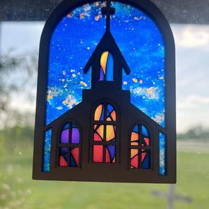 May include: Wooden church-shaped ornament with a stained-glass effect. The ornament features a cross on top and colorful windows in shades of purple, orange, and red. The background is a vibrant blue, resembling a sky.