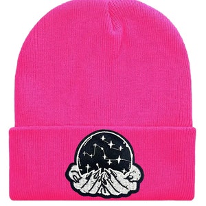 May include: A pink knit beanie with a black and white embroidered patch of a hand holding a crystal ball with stars inside.