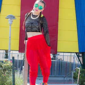 May include: A woman wearing a black mesh crop top, red trousers, and rainbow platform trainers. She is standing in front of a yellow and red building.