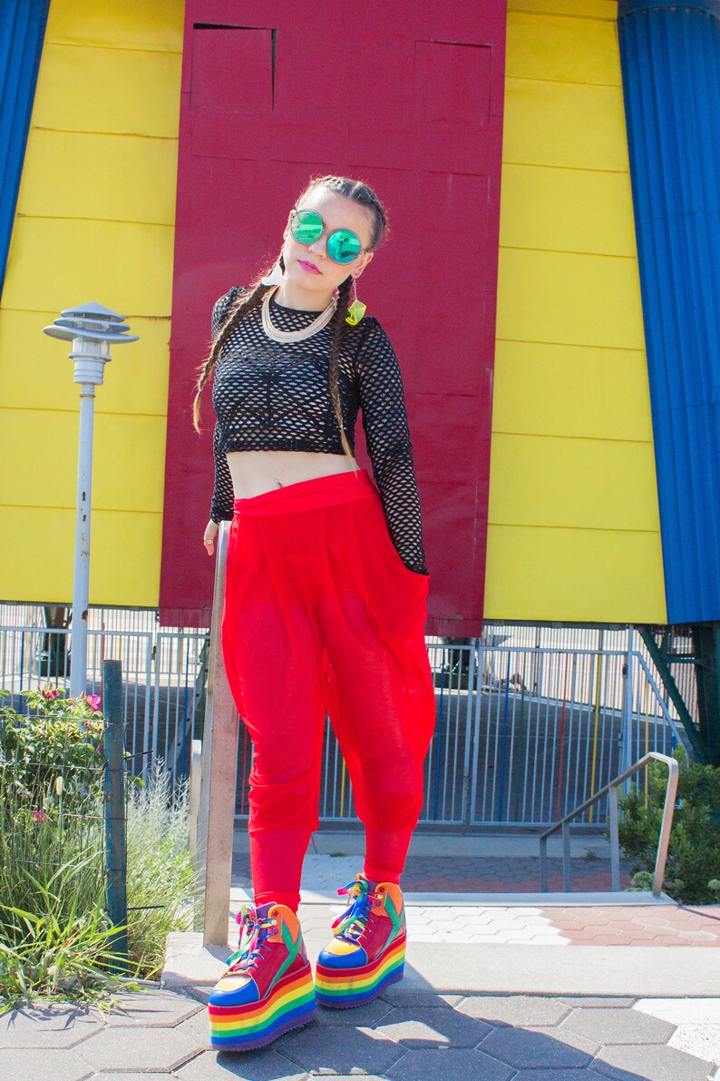 May include: A woman wearing a black mesh crop top, red trousers, and rainbow platform trainers. She is standing in front of a yellow and red building.