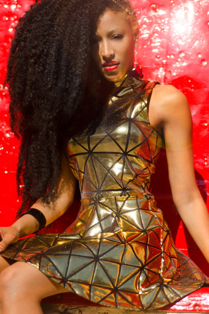 May include: A woman with long black hair wearing a gold metallic sleeveless dress with a geometric pattern. The dress has a high neckline and a fitted bodice. The woman is sitting on a red surface.