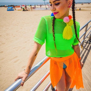 May include: A woman wearing a bright green mesh crop top and an orange mesh skirt. She is standing on a boardwalk with a sandy beach in the background. She has her hair in two braids with colorful pom-pom earrings.