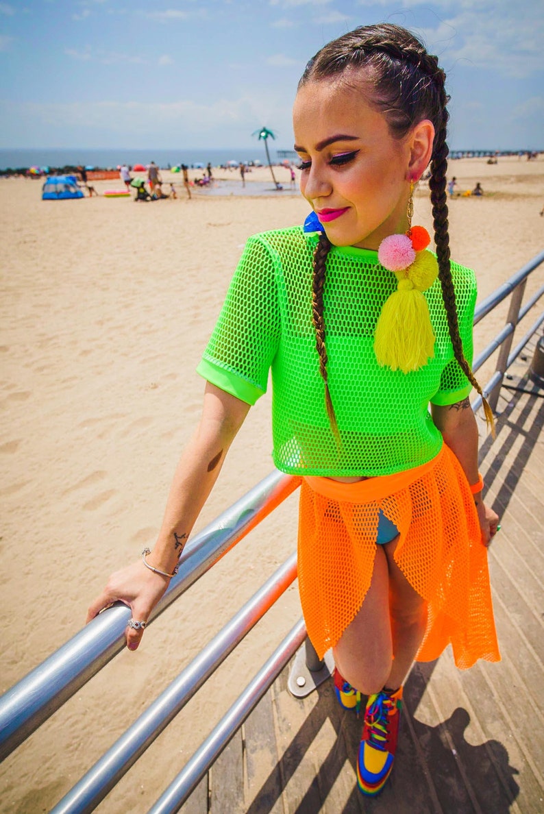 May include: A woman wearing a bright green mesh crop top and an orange mesh skirt. She is standing on a boardwalk with a sandy beach in the background. She has her hair in two braids with colorful pom-pom earrings.