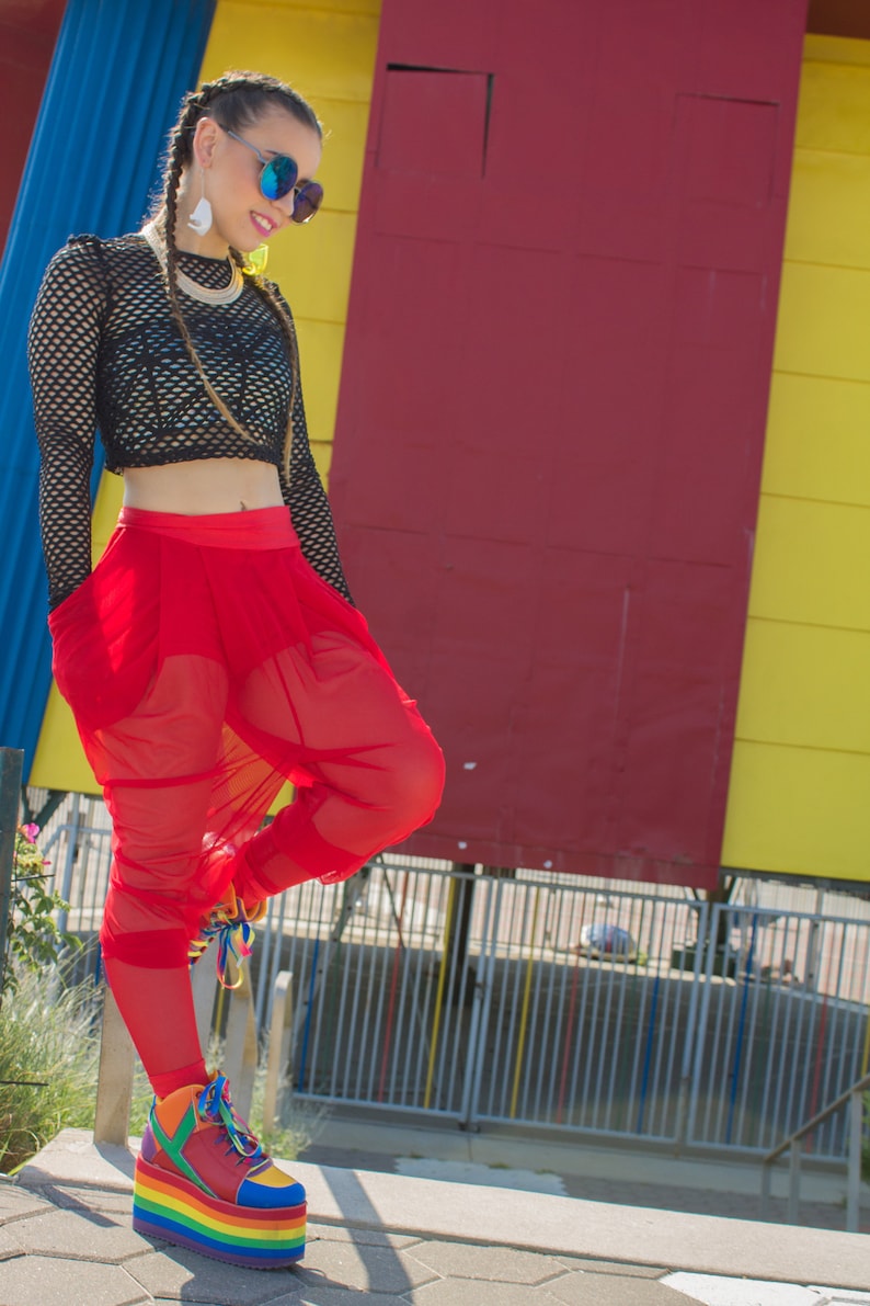 May include: A woman wearing a black fishnet top, red sheer trousers, and rainbow platform trainers. She is standing in front of a colourful wall.