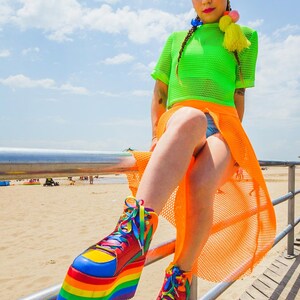May include: A person wearing a bright green mesh top and an orange mesh skirt, sitting on a metal railing. They are wearing rainbow platform shoes with yellow, red, blue, green, and purple accents.