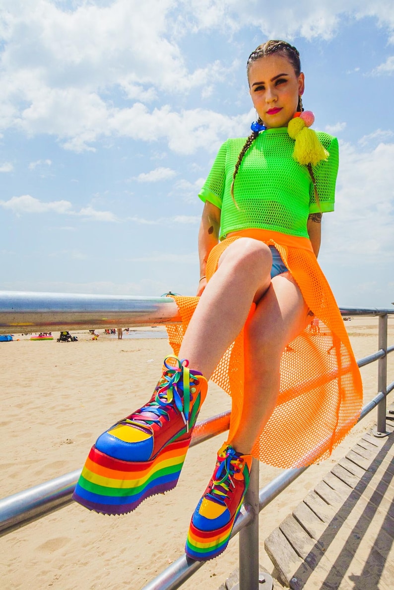 May include: A person wearing a bright green mesh top and an orange mesh skirt, sitting on a metal railing. They are wearing rainbow platform shoes with yellow, red, blue, green, and purple accents.